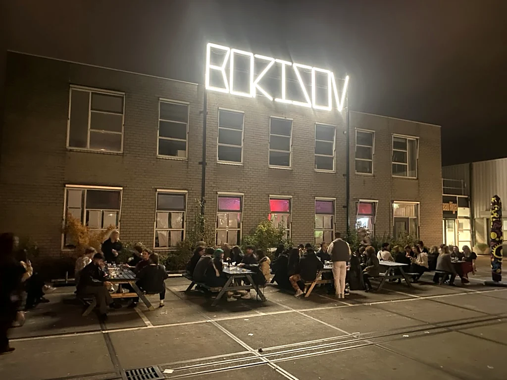 Het gebouw met het lichtbord aan, tafels vol op het plein, Borisov Amsterdam